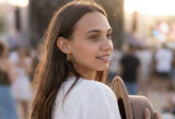 A woman with long brown hair wearing a white top and gold hoop earrings, holding a brown hat in a blurred outdoor crowd setting. A close-up portrait of a woman wearing the elegant Alba earrings, a gold link earring by Delani Jewelry and a gold chain necklace, set against a blurred outdoor background.