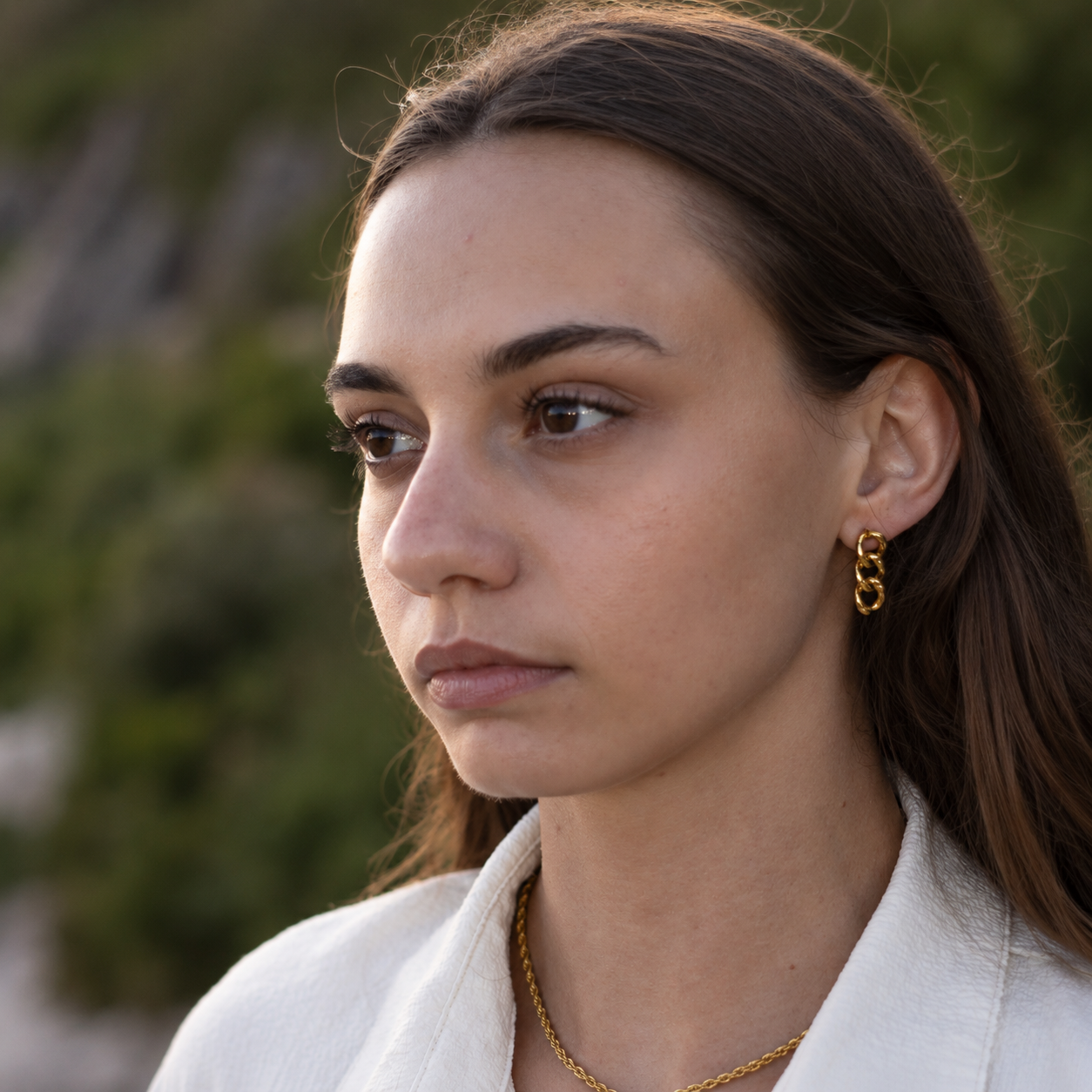 A close-up portrait of a woman wearing the elegant Alba earrings, a gold link earring by Delani Jewelry and a gold chain necklace, set against a blurred outdoor background.