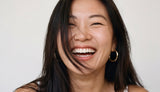 Woman with dark hair and gold Holly Hoop earrings smiling against a plain background from Delani Jewelry