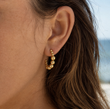 A close-up of a person's ear wearing a small gold hoop earring with a row of tiny gold beads along the front, set against a softly blurred outdoor background. The design is gold and looks like bubbles in ascending sizes from the center. These are the Kendall Earrings with that are waterproof, tarnishproof with a lifetime color guarantee jewelry from Delani Jewelry.
