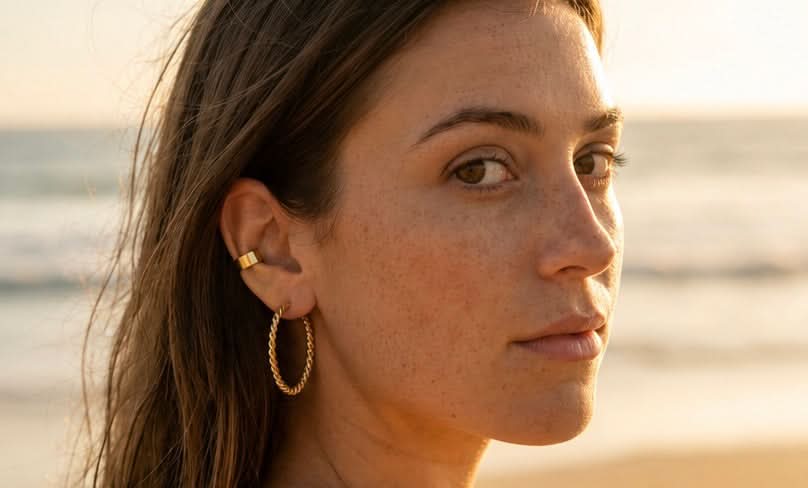 A close-up portrait of a woman with brown hair wearing a small gold hoop earring and a gold ear cuff, photographed outdoors against a softly blurred beach background at sunset.
