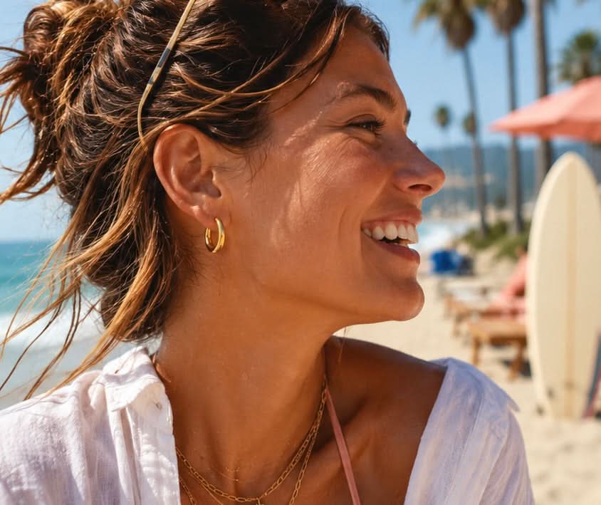 Model wearing Delani Jewelry gold huggie hoop earrings at the beach, showcasing effortless coastal style with jewelry made to swim, shower, and live in. Waterproof, tarnish-proof, hypoallergenic, and backed by our Lifetime Color Guarantee.