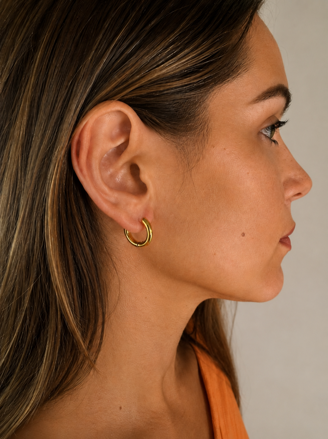 Close-up of Myra Earrings by Delani Jewelry, a woman wearing a gold hoop earring against a neutral background