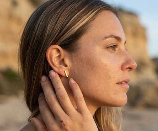 Woman wearing Delani Jewelry gold bar stud earrings at the beach, featuring sleek minimalist rectangular studs with a polished finish for effortless everyday style and modern ear stacking. Named Namaari Studs