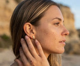 Woman wearing Delani Jewelry gold bar stud earrings at the beach, featuring sleek minimalist rectangular studs with a polished finish for effortless everyday style and modern ear stacking. Named Namaari Studs