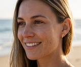 Woman wearing Delani Jewelry gold bar stud earrings on the beach, showcasing minimalist rectangular earrings with a sleek polished finish, perfect for first, second, or third lobe piercings and everyday styling. Named Namaari Studs