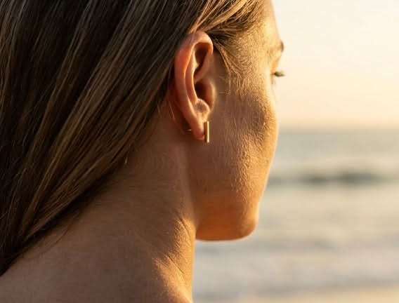 Woman wearing Delani Jewelry gold bar stud earrings at the beach, featuring sleek minimalist rectangular studs for effortless everyday style and modern ear stacking. Named Namaari Studs