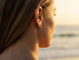 Woman wearing Delani Jewelry gold bar stud earrings at the beach, featuring sleek minimalist rectangular studs for effortless everyday style and modern ear stacking. Named Namaari Studs