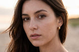 A close-up portrait of a woman with dark brown hair and freckles, wearing a small gold hoop earring, set against a softly blurred beach background at sunset. Delani gold twisted stud earrings on a beach at golden hour. Waterproof, tarnish-proof earrings with a lifetime color guarantee, showcased in a luxury lifestyle portrait.