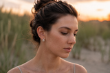 A woman with her hair in a messy bun wearing a small silver earring, photographed outdoors in warm sunset light with blurred grassy dunes in the background. Delani silver twisted stud earrings on a beach at golden hour. Waterproof, tarnish-proof earrings with a lifetime color guarantee, showcased in a luxury lifestyle portrait.