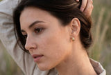 A close-up profile shot of a woman wearing a small gold textured hoop earring, with her hand in her hair, set against a soft-focus natural background. wearing Delani gold twisted stud earrings on a beach at golden hour. Waterproof, tarnish-proof earrings with a lifetime color guarantee, showcased in a luxury lifestyle portrait and effortless coastal style