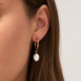 A close-up of a model's ear wearing a gold huggie hoop earring with a dangling white baroque pearl. Delani Jewelry's Pola Hoops gold freshwater pearl drop hoop earrings with organic baroque pearls. Waterproof, tarnish-proof, hypoallergenic earrings backed by a Lifetime Color Guarantee.