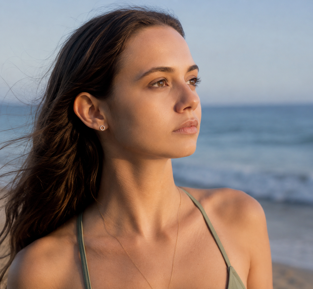 A woman with long dark brown hair stands on a beach, wearing a thin olive green halter top and delicate gold-toned jewelry, with the ocean and sky blurred in the background. Delani Jewelry's Rana earrings in Gold. Earrings are waterproof, tarnish-proof and have a lifetime color guarantee. 