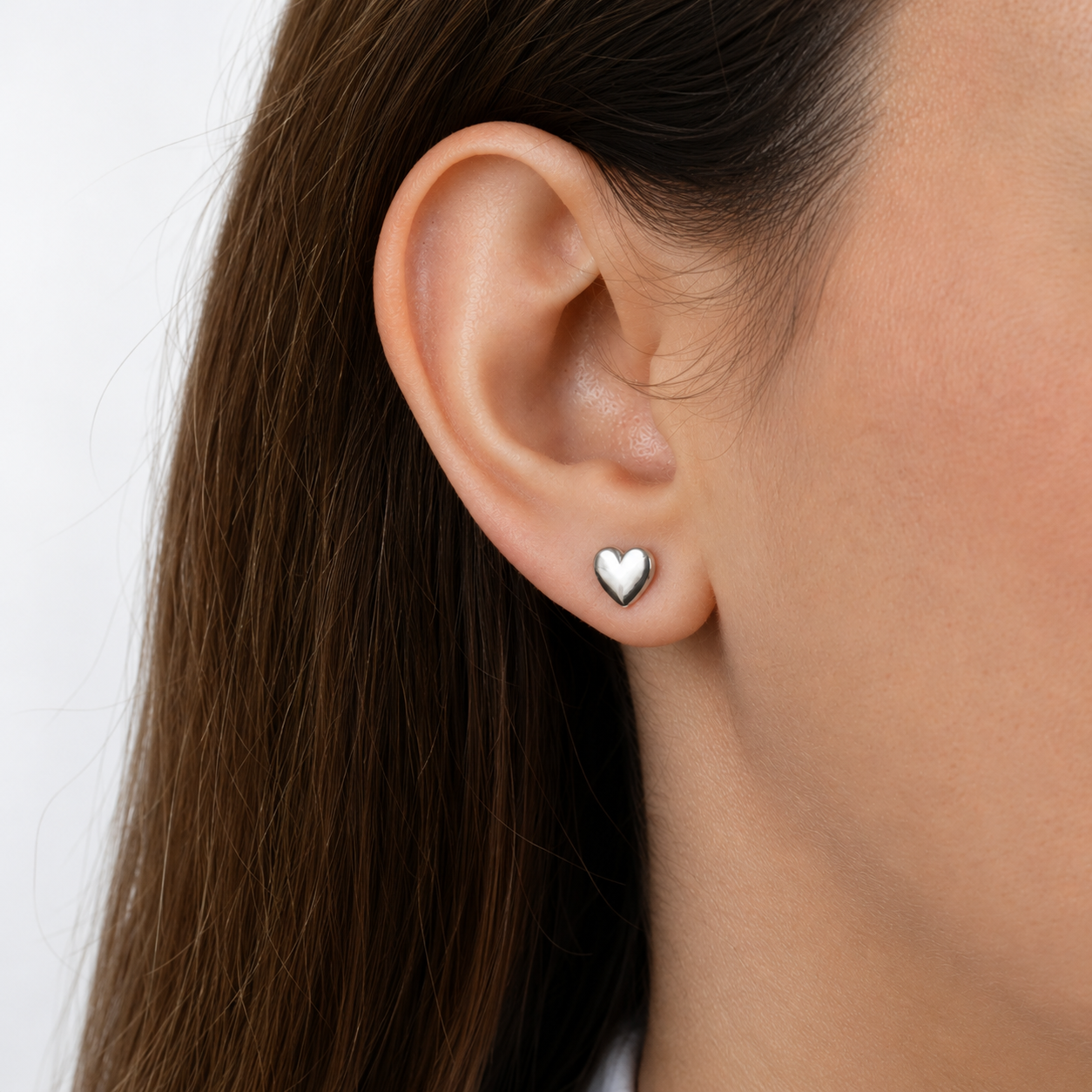 A close-up of a woman's ear wearing a small polished silver heart stud earring against a clean white background. Minimalist silver heart stud earrings by Delani Jewelry, waterproof, tarnish-proof stainless steel with a Lifetime Color Guarantee, shown in a close-up ear styling.