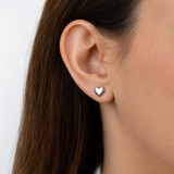 A close-up of a woman's ear wearing a small polished silver heart stud earring against a clean white background. Minimalist silver heart stud earrings by Delani Jewelry, waterproof, tarnish-proof stainless steel with a Lifetime Color Guarantee, shown in a close-up ear styling.