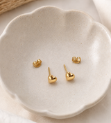 A pair of small gold heart stud earrings displayed on a scalloped off-white ceramic dish. Minimalist gold heart stud earrings by Delani Jewelry, waterproof, tarnish-proof stainless steel with a Lifetime Color Guarantee, shown in a close-up ear styling.