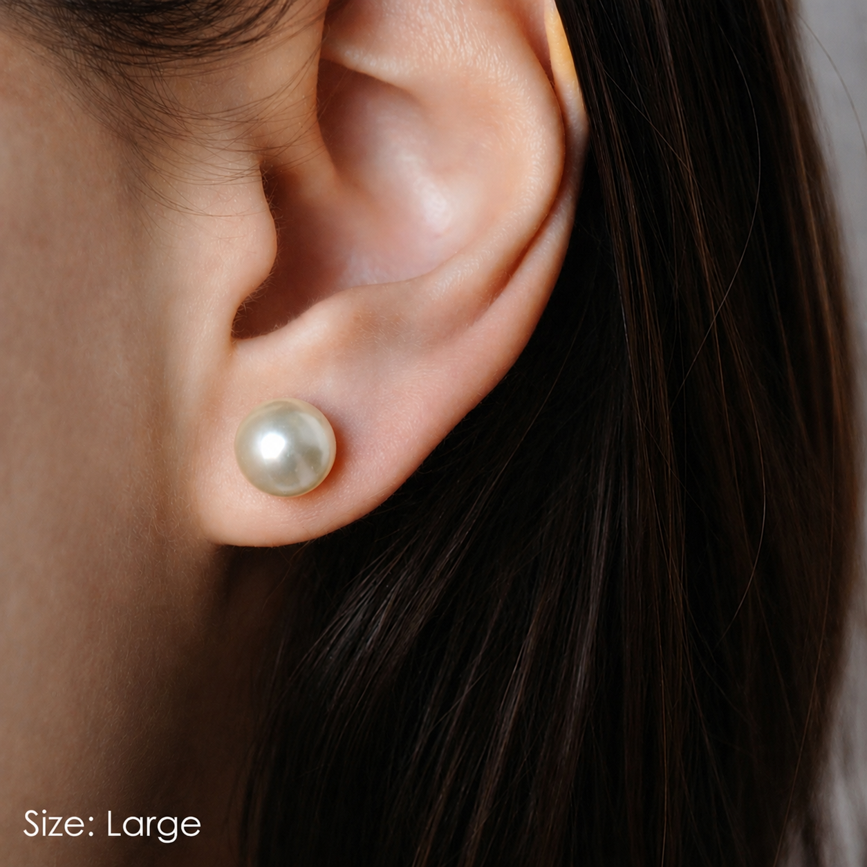 Close-up of an ear wearing a pearl earring with dark hair partially visible. Size Large. Classic Satya pearl earrings from Delani Jewelry with luminous white pearls, stainless steel posts, waterproof and tarnish-proof design, perfect for everyday wear with a lifetime color guarantee.