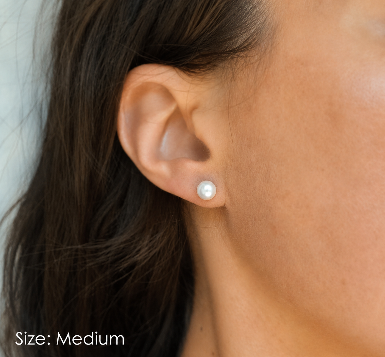 A close-up of a person's ear wearing a single round white pearl stud earring, with the text 'Size: Medium' in the lower-left corner. Classic Satya pearl earrings from Delani Jewelry with luminous white pearls, stainless steel posts, waterproof and tarnish-proof design, perfect for everyday wear with a lifetime color guarantee.