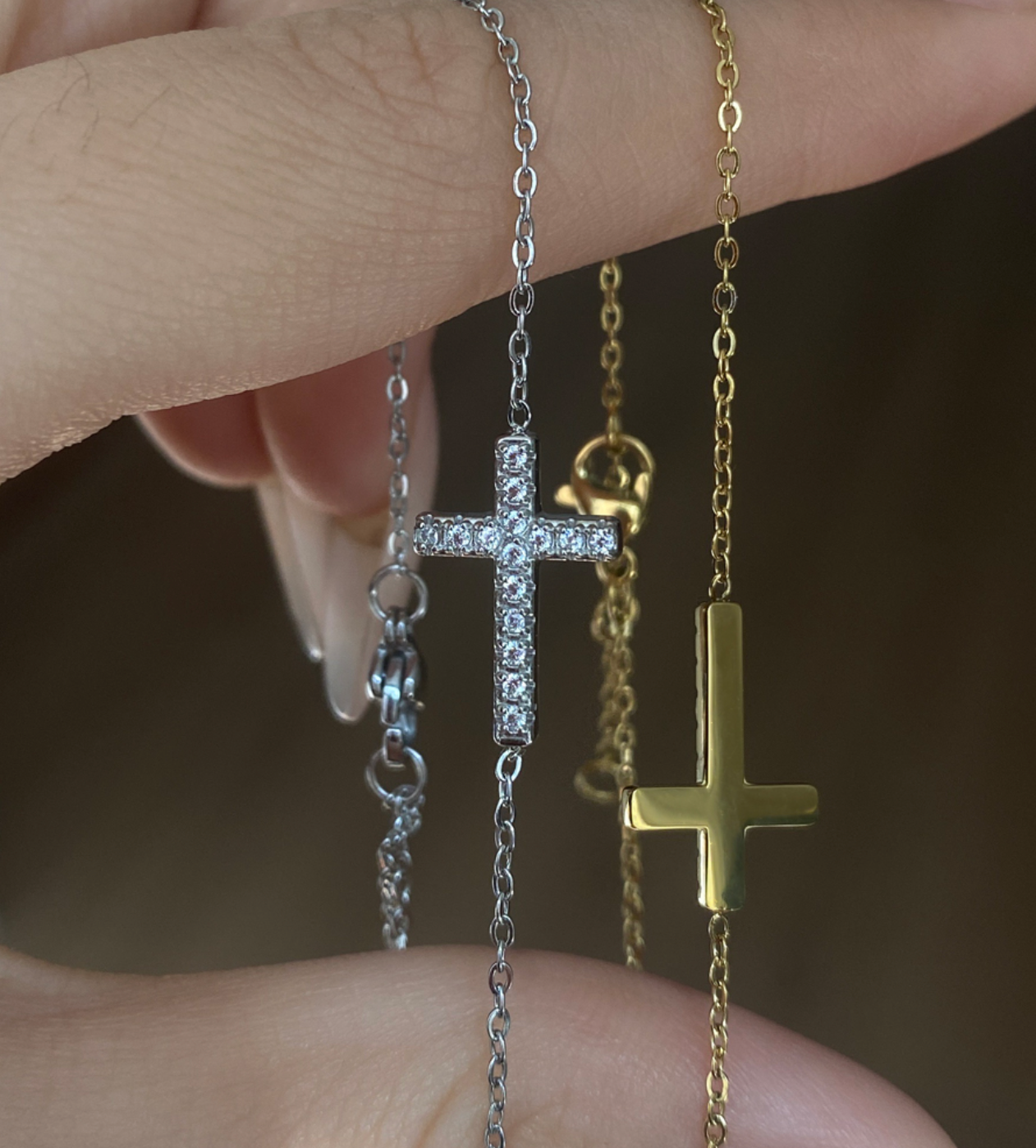 Two necklaces with cross pendants, one silver and one gold, held against a neutral background to showcase color choices. From Delani Jewelry