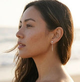 Model wearing Delani Jewelry's gold Shyla chain drop earrings with a bezel-set stone and delicate chain. Waterproof, tarnish-proof, hypoallergenic earrings backed by a lifetime guarantee. Mismatched stones numbers create a drama and appeal unique to the Shyla Earrings. 